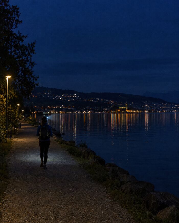 Ein einzelner Wanderer mit Stirnlampe nachts auf dem Uferweg direkt am See, von hinten